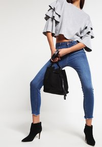 Grauer, kurzer Sweatshirt mit Rüschensleeves, kombiniert mit blauen Skinny Jeans und schwarzen Ankle Boots. Schwarze Bucket Bag in der Hand gehalten.