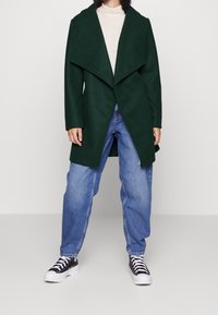 Grön ullblandad kappa med en bred, öppen krage och asymmetrisk front, kombinerad med ljusblå denimjeans och svartvita sneakers.