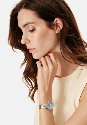 Sølvfarvet armbåndsur med rund, lyseblå urskive og krystal accenter; har en kædet metalrem og et minimalistisk design.