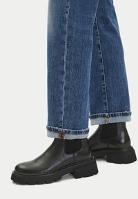 Stopy w czarnych, skórzanych botkach Chelsea na masywnej podeszwie, zestawione z podwiniętymi niebieskimi dżinsami na białym, jednolitym tle.