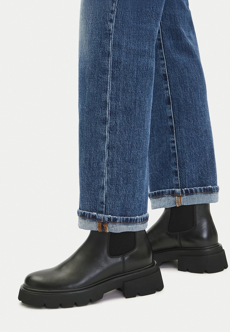 Stopy w czarnych, skórzanych botkach Chelsea na masywnej podeszwie, zestawione z podwiniętymi niebieskimi dżinsami na białym, jednolitym tle.