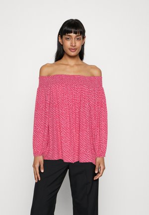 Ružová blúzka s odhalenými ramenami a štruktúrovaným vzorom, dlhé rukávy a voľný strih, skombinovaná s tmavými nohavicami.