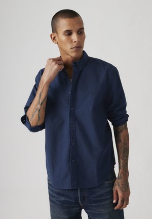Jeune homme aux cheveux courts portant une chemise bleu marine boutonnée avec les manches retroussées et un jean bleu, montrant des tatouages sur les deux avant-bras.