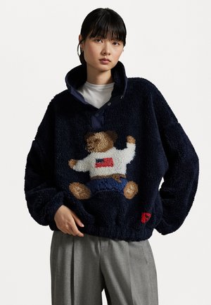 Pullover in pile navy con design di orsetto; presenta un colletto alto, chiusura a bottoni e tessuto testurizzato. Sono presenti dettagli in rosso e bianco.