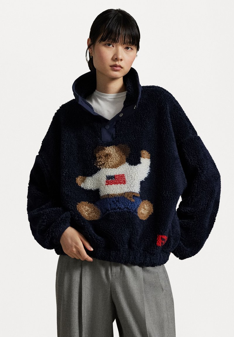 Marinefarbener Fleece-Pullover mit einem Teddy-Bären-Design; verfügt über einen hohen Kragen, einen Knopfverschluss und einen strukturierten Stoff. Rote und weiße Akzente sind vorhanden.