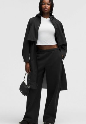 Mujer con un abrigo negro largo, pantalones anchos negros, top corto blanco, sosteniendo un pequeño bolso negro, de pie frente a un fondo liso.