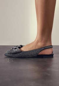 Juodos slingback tipo plokščiapadžiai batai su kristalų apdaila, maža lankeliu prie piršto, lygiu paviršiumi ir žemu, plačiu padu.