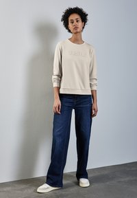 Ljusbeige sweatshirt med präglad text "SUBTLE", i kombination med mörkblå vida jeans och krämfärgade sneakers, mot en neutral bakgrund.
