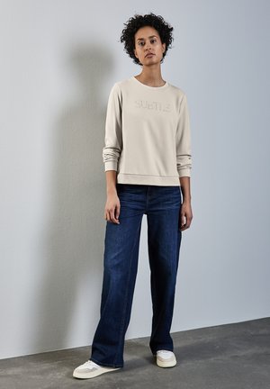 Felpa beige chiaro con testo in rilievo "SUBTLE", abbinata a jeans a gamba larga blu scuro e sneakers color crema, su uno sfondo neutro.