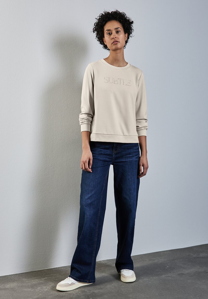 Ljusbeige sweatshirt med präglad text "SUBTLE", i kombination med mörkblå vida jeans och krämfärgade sneakers, mot en neutral bakgrund.
