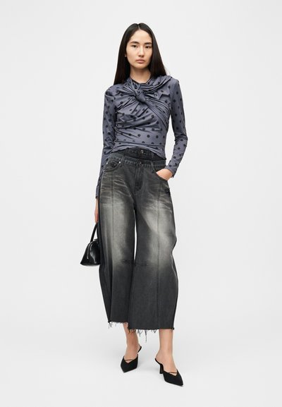 Μπλούζα με μακριά μανίκια, γκρί με πουά, με λεπτομέρεια δέσιμου στον κόμπο, συνδυασμένη με φαρδιά, σκισμένα μαύρα τζιν. Μαύρη τσάντα χειρός και μυτερά φλατ παπούτσια ολοκληρώνουν το look.