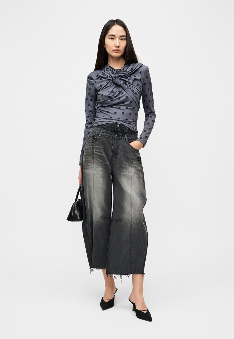 Haut à manches longues gris à pois avec détail noué, associé à un jean noir large et déchiré. Sac à main noir et ballerines pointues complètent le look.