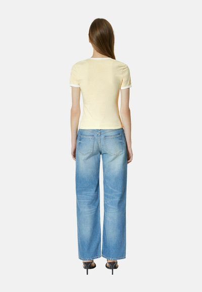 Woman standing facing away wearing a pale yellow short-sleeve shirt, light blue wide-leg jeans, and black heeled sandals.