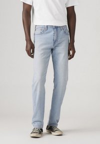 Világoskék denim farmernadrág egyenes lábbal, öt zsebes stílusban, és látható varrásokkal. Fekete sportcipővel párosítva, mely kopott és texturált.