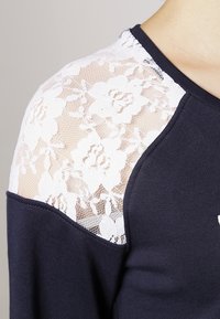 Maglione blu con corpo in maglia texture, caratterizzato da pannelli di pizzo bianco sulle spalle con motivi floreali, scollatura rotonda e polsini a costine.