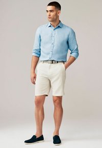 Cămașă din in albastru deschis cu nasturi, pantaloni scurți crem, curea gri cu model și pantofi slip-on de culoare albastră închisă. Texturi fine cu o potrivire casuală.