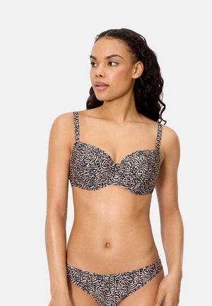 Mujer con bikini estampado en negro y beige con top con aros, mirando hacia la izquierda sobre un fondo liso claro.