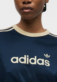Detail osoby nosící sportovní tričko Adidas tmavě modré barvy s béžovým lemováním a logem uprostřed hrudi.