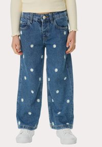 Jeans de tiro ancho de mezclilla con bordado de margaritas blancas, cinco bolsillos y cierre de botón. Tejido azul con un acabado de lavado medio. Zapatillas blancas.