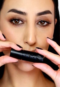 Tube de rouge à lèvres noir tenu entre des doigts, avec du texte en relief. Le modèle a un maquillage neutre et des ongles manucurés foncés. Gros plan sur le produit.