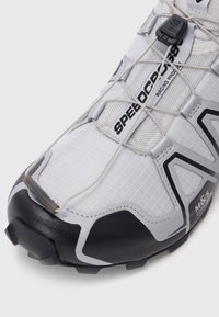 Tênis athletic brancos com parte superior em tecido texturizado, biqueira de borracha preta, cordões elásticos e detalhes em prata com um design estilo corrida.