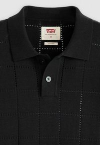 Polo negro con un patrón texturizado, que cuenta con una tapeta de dos botones y un botón azul. La etiqueta dice "Levi's, Standard, M."