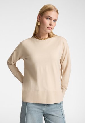 Modèle féminin portant un pull beige à manches longues et un jean bleu clair, accessoirisé d'un collier chaîne en or et de boucles d'oreilles, sur fond blanc.