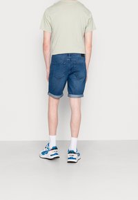 Shorts en denim avec un ourlet retroussé, de couleur bleu moyen, et deux poches arrière. Assortis avec un t-shirt vert clair et des baskets blanches avec des accents bleus.