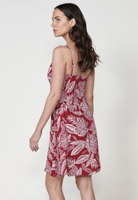 Rotes Blumen-Sommerkleid mit weißen Blattmustern, dünnen Trägern und einem taillierten Oberteil, das in einen Rock ausläuft, aus leichtem Stoff gefertigt.