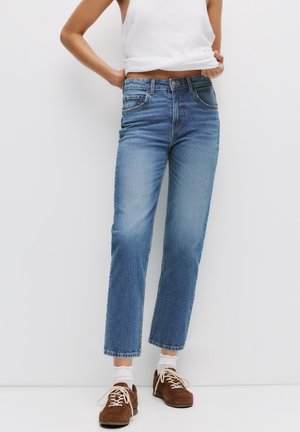 Personne portant un jean bleu droit, des chaussettes blanches, des chaussures marron à lacets et un haut blanc sans manches, debout devant un fond blanc uni.