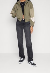 Woman wearing a cropped olive green jacket, black crop top, high-waisted dark gray jeans, and black-and-white sneakers, standing against white background.