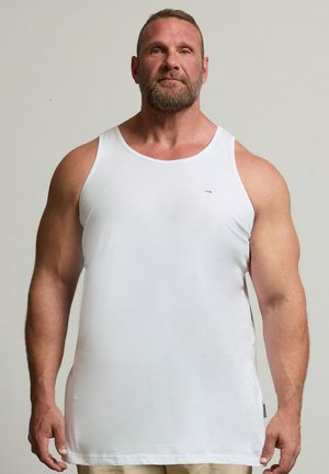Muskulöser Mann mit Bart, der ein weißes Tanktop und eine helle Hose trägt, steht frontal vor einem schlichten, hellen Hintergrund.