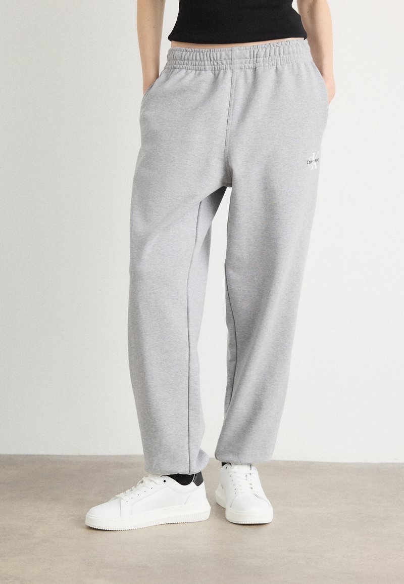 Lichtgrijze sweatpants met een elastische tailleband, rechte broekspijpen en een klein logo detail. Gekocht met witte sneakers op een neutrale vloer.
