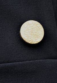 Czarna tkanina o textured surface, z okrągłym, metalicznym złotym guzikiem, który ma liniowy wzór. Guzik dodaje kontrastujący detal.