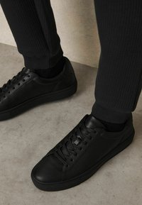 Zapatillas negras de cuero con cordones planos, puntera redonda y textura suave. Combinadas con pantalones negros de canalé, parcialmente visibles arriba.
