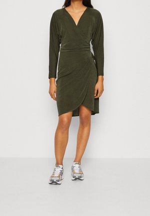 Kvinde iført olivengrøn langærmet slå-om kjole kombineret med hvide og grå atletiksneakers, stående foran en ensfarvet baggrund.