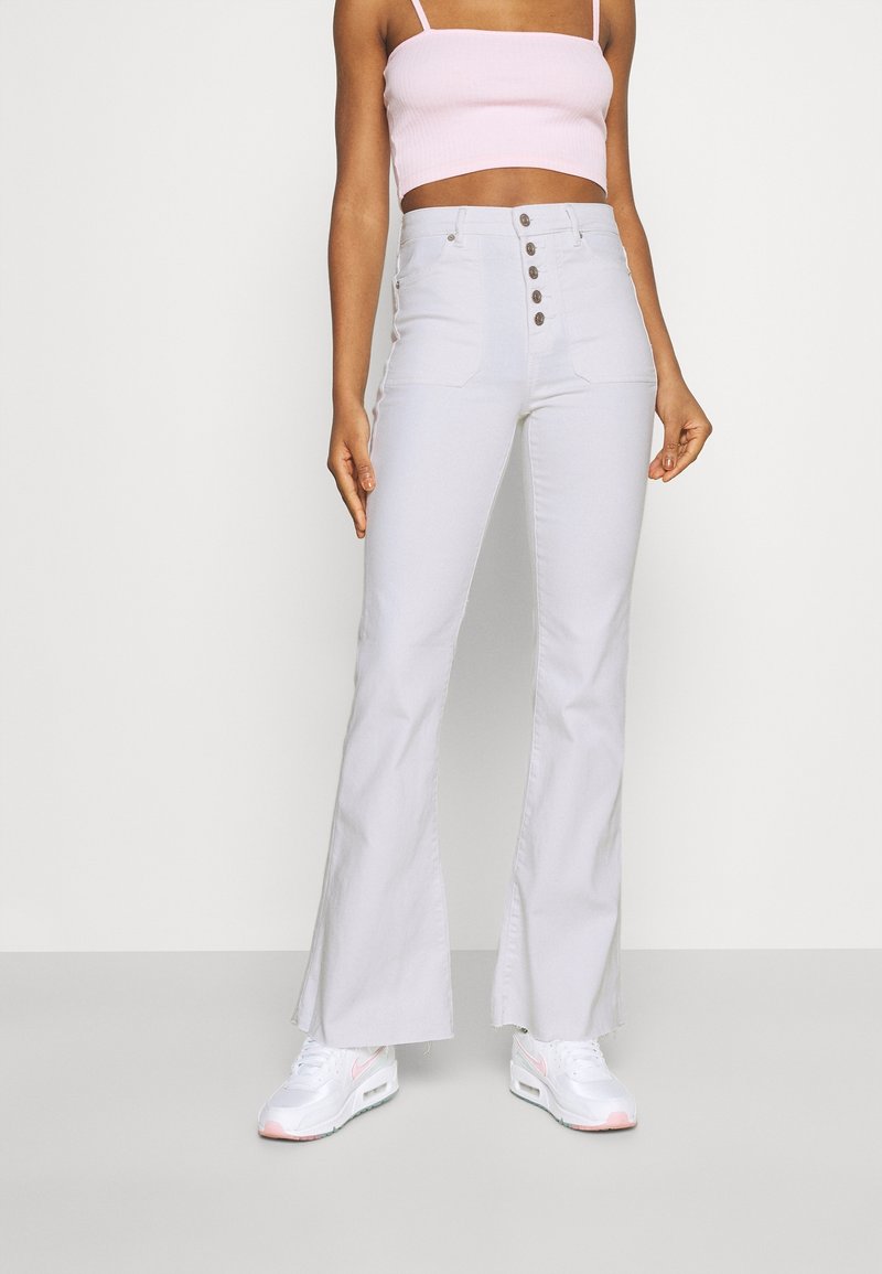 Femme portant un jean évasé taille haute blanc avec des boutons sur le devant, un débardeur court rose clair et des baskets blanches avec des détails roses.