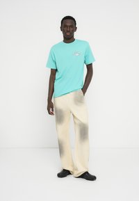 Τυρκουάζ βαμβακερό μπλουζάκι με γραφικό λογότυπο, σετ με κρεμ παντελόνι με ξεθωριασμένο γκρι μοτίβο και μαύρα παπούτσια.