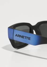 Černé sluneční brýle s matným povrchem a modrými gradientními stranicemi. Na boku se nachází výrazné logo "ARNETTE". Skla jsou tmavě zabarvená.