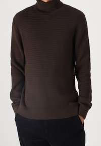 Pull à col roulé marron foncé avec un motif côtelé texturé, doté de manches longues et d'une coupe décontractée. Le matériau présente une apparence douce et en maille.
