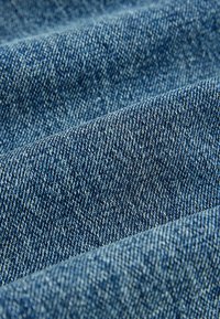 Gros plan sur un tissu en denim bleu avec un tissage texturé visible et de légers plis formant des lignes diagonales.