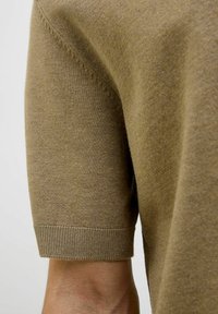 Trumpas rankovėmis rudas megztinis, pasižymintis glotnia tekstūra, jautriomis rankogaliais ir subtiliais siuvinėjimo akcentais neutralioje fone.