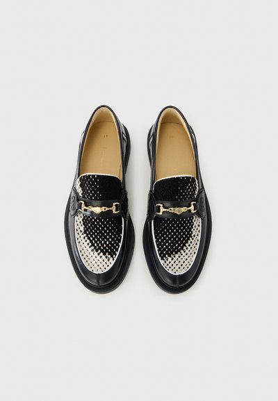 Black and white loafers featuring a textured upper with a dotted pattern, leather accents, a gold-toned clasp, and a rounded toe design.
