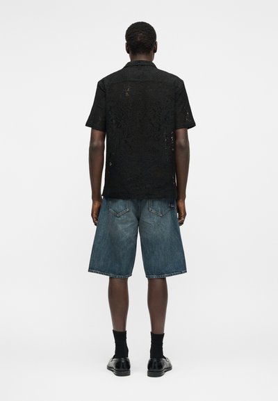 Άνδρας που στέκεται με την πλάτη προς τα πίσω, φοράει μαύρο δαντελωτό πουκάμισο με κοντά μανίκια, μπλε τζιν σορτς μέχρι το γόνατο, μαύρες κάλτσες και μαύρα επίσημα παπούτσια.