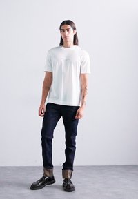 Young person with long hair stands against plain white wall wearing white graphic t-shirt, black jeans with folded cuffs, and black shoes.