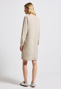 Nainen, jolla on vaaleat hiukset, pukeutuneena polvimittaseen beigeen mekkoon ja valkoisiin lenkkareihin, seisoo selkä cameraan päin yksinkertaista taustaa vasten.