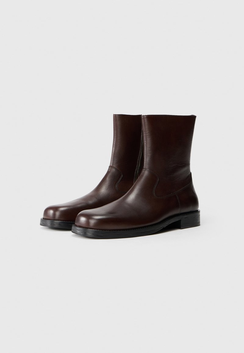 Bottines en cuir marron avec une texture lisse, bout arrondi, couture minimale et semelle en caoutchouc noir. Elles sont équipées d'une fermeture éclair sur le côté.