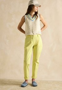 Kreminės spalvos bepevės megztinės su keturių raštų ir mėlynu apvadu, derinamos su šviesiai geltonomis kelnėmis ir mėlynais zomšiniais bateliais.