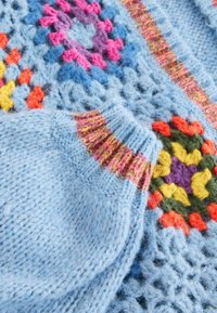Moufles tricotées bleu clair avec poignets côtelés multicolores posées sur une couverture bleue au crochet ornée de motifs floraux éclatants.