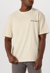T-shirt in cotone beige con vestibilità rilassata. Presenta un collo rotondo e maniche corte. Logo "ANERKJENDT" stampato sulla parte superiore sinistra del petto.